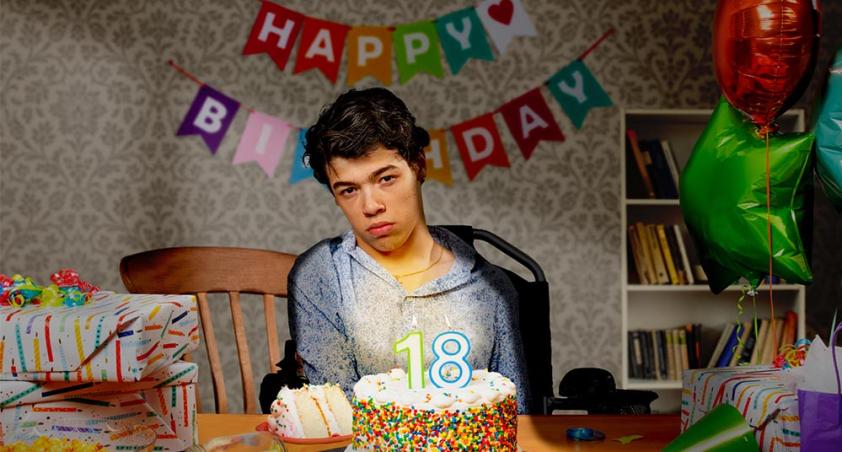 A teenage boy, who uses a wheelchair, sitting at a table in front of a birthday cake with lit ‘18’ candles.